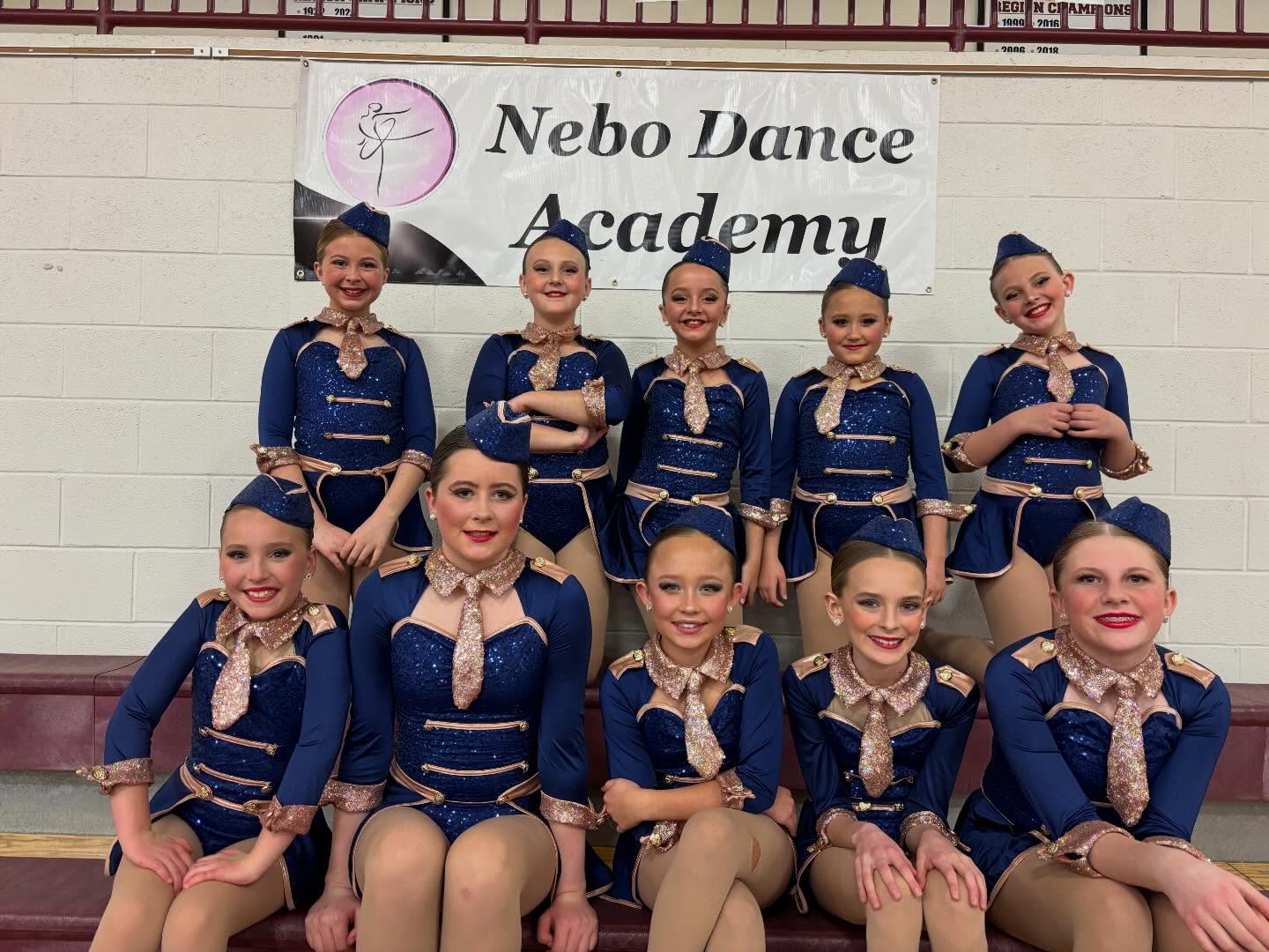 Nebo Dance Academy team in navy costumes posing under the studio banner